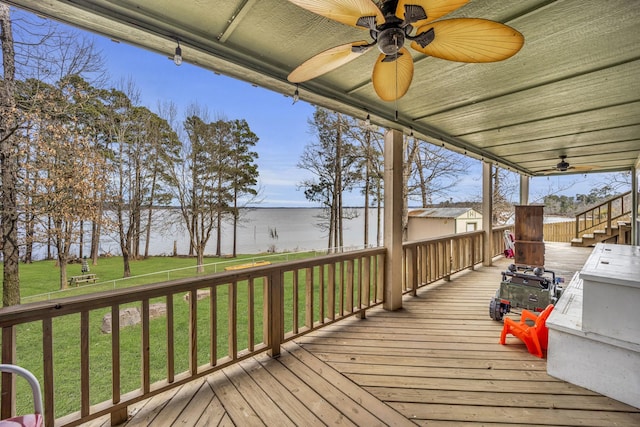 wooden deck featuring a water view, a ceiling fan, a lawn, and an outdoor structure