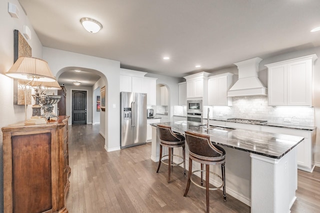 kitchen with arched walkways, wood finished floors, stainless steel appliances, premium range hood, and a sink