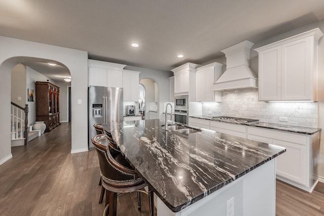 kitchen with tasteful backsplash, arched walkways, stainless steel appliances, a large island with sink, and premium range hood