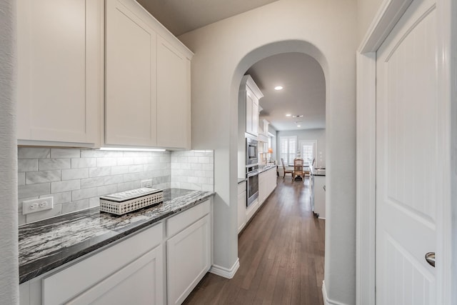 kitchen with arched walkways, dark wood-style flooring, decorative backsplash, white cabinets, and dark stone countertops