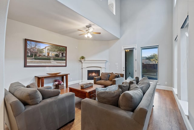 living area featuring a towering ceiling, a glass covered fireplace, ceiling fan, wood finished floors, and baseboards