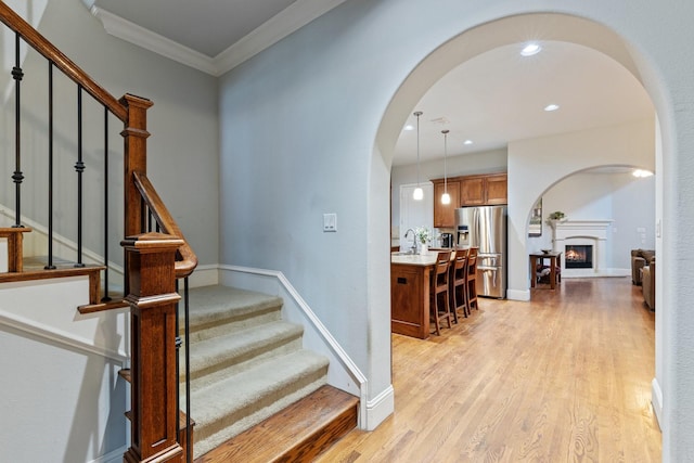 staircase featuring arched walkways, a warm lit fireplace, wood finished floors, baseboards, and crown molding