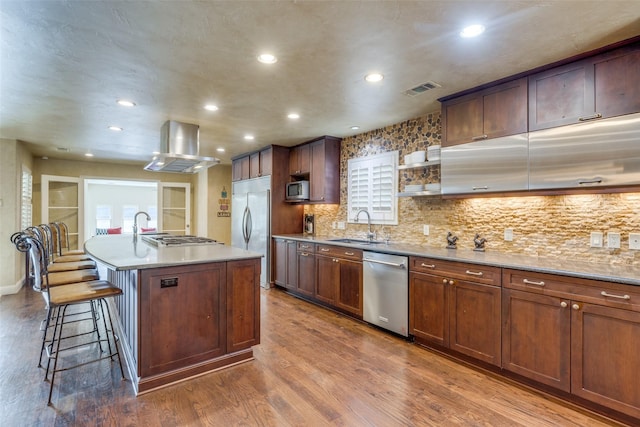 kitchen with a center island with sink, appliances with stainless steel finishes, dark wood-style flooring, island exhaust hood, and a sink