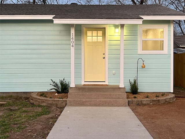 view of exterior entry with a shingled roof and fence