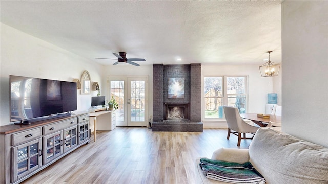 living area with french doors, a brick fireplace, a textured ceiling, light wood-type flooring, and ceiling fan with notable chandelier