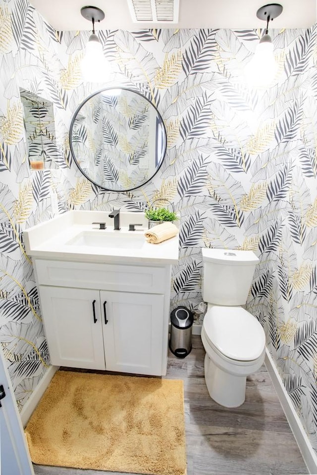 half bath featuring toilet, wood finished floors, vanity, baseboards, and wallpapered walls