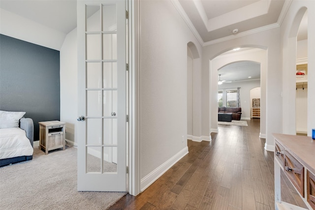 corridor featuring baseboards, arched walkways, a raised ceiling, wood finished floors, and crown molding