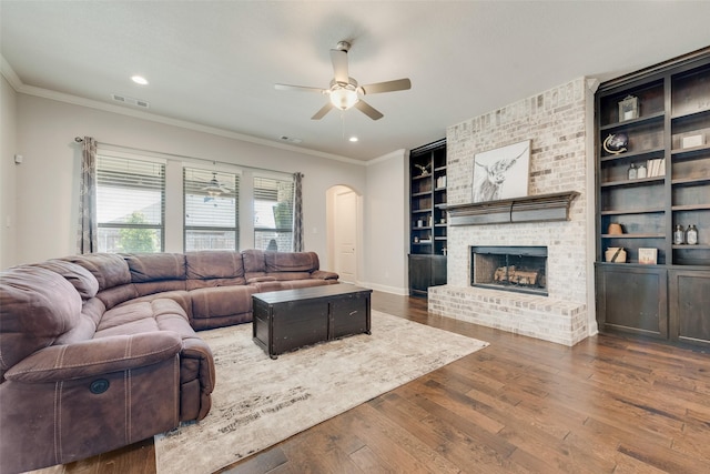 living room with arched walkways, a fireplace, ornamental molding, a ceiling fan, and wood finished floors