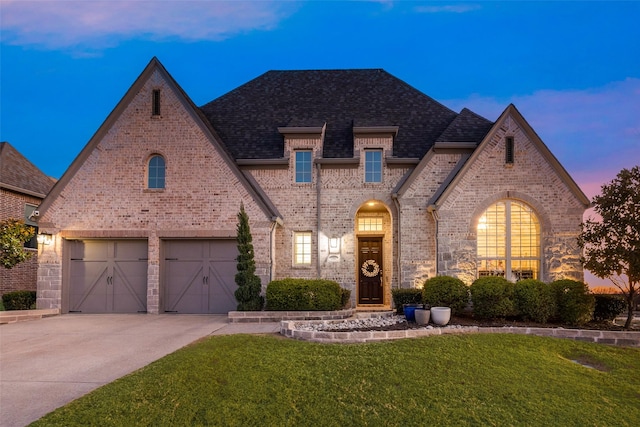 french country inspired facade with brick siding, concrete driveway, a yard, and a shingled roof