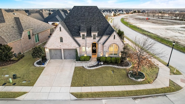 french country inspired facade featuring a garage, driveway, roof with shingles, and a front yard