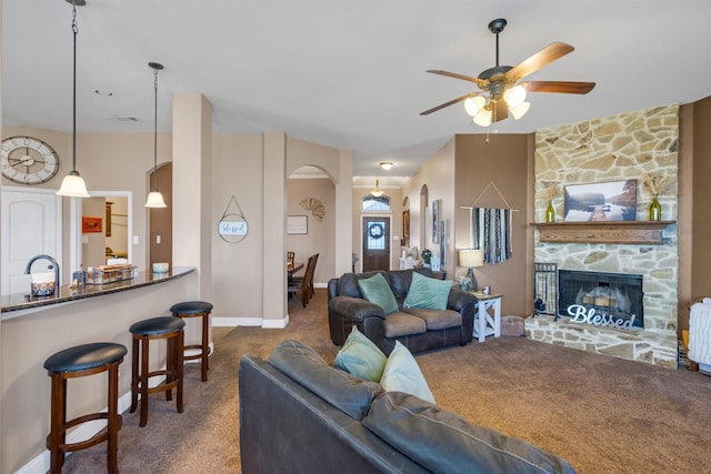 living room featuring carpet floors, arched walkways, a ceiling fan, a stone fireplace, and baseboards