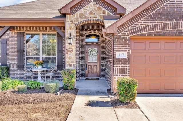 entrance to property featuring a garage, stone siding, brick siding, and roof with shingles