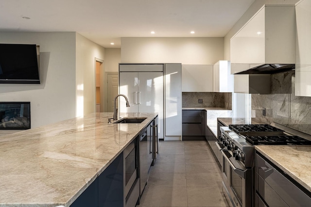 kitchen featuring high end appliances, modern cabinets, light stone countertops, wall chimney range hood, and a sink