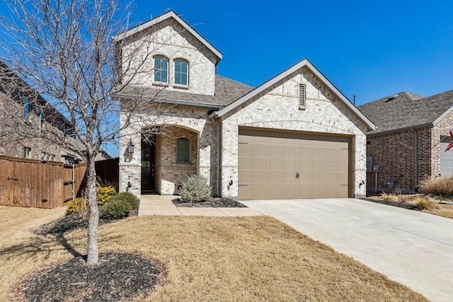 french country style house with a garage, a shingled roof, concrete driveway, fence, and brick siding