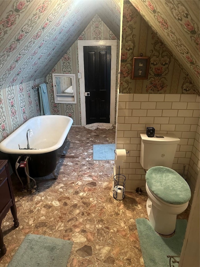 bathroom with lofted ceiling, toilet, wainscoting, a soaking tub, and wallpapered walls