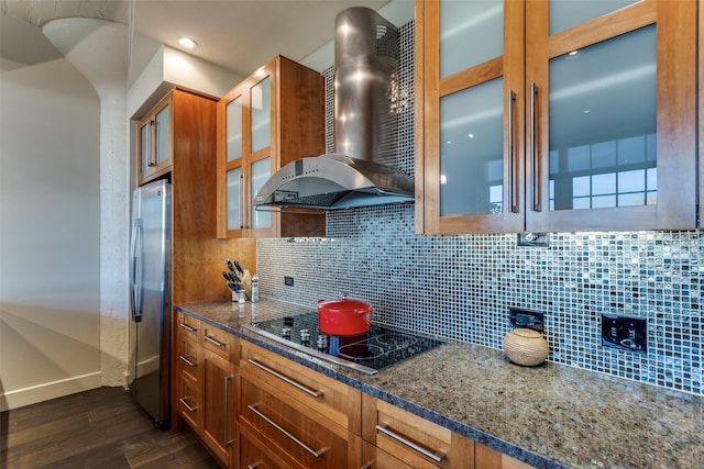 kitchen featuring dark stone countertops, freestanding refrigerator, black electric cooktop, wall chimney range hood, and backsplash