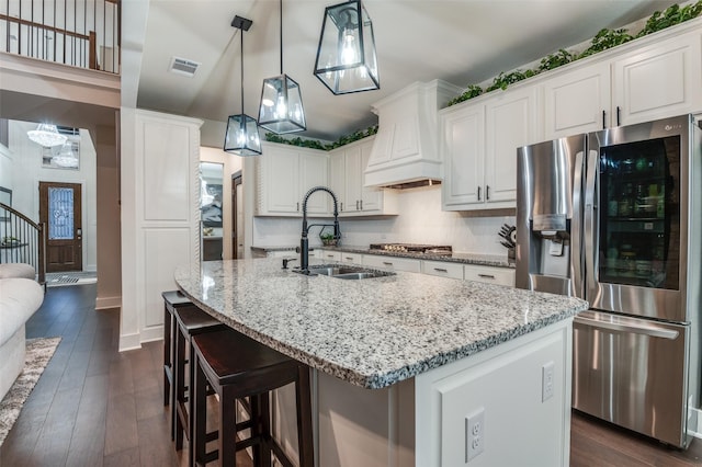 kitchen with light stone counters, a kitchen island with sink, a sink, appliances with stainless steel finishes, and custom range hood