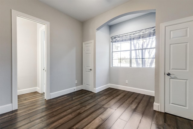 unfurnished bedroom featuring arched walkways, dark wood finished floors, and baseboards