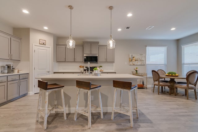 kitchen featuring stainless steel microwave, a kitchen breakfast bar, a kitchen island with sink, light countertops, and gray cabinetry