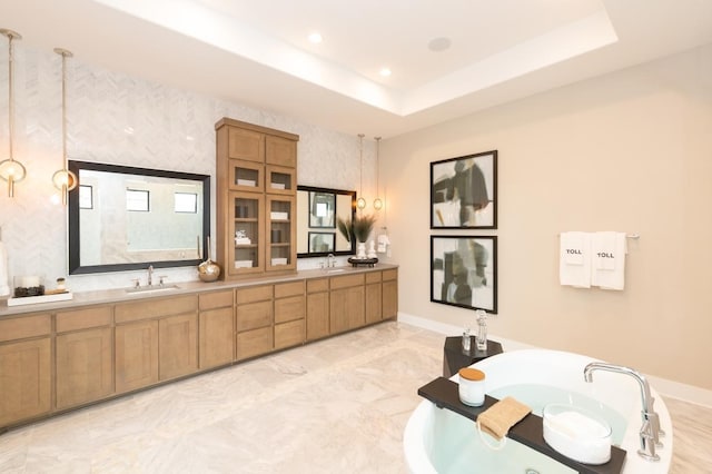 bathroom featuring baseboards, a tray ceiling, a freestanding bath, a sink, and recessed lighting