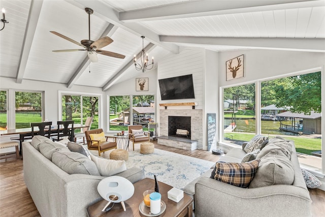 living room featuring ceiling fan with notable chandelier, beamed ceiling, a stone fireplace, and wood finished floors