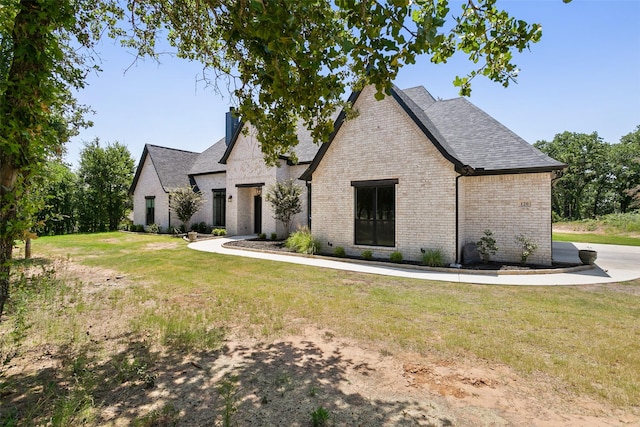 french country style house featuring roof with shingles, brick siding, a chimney, and a front yard