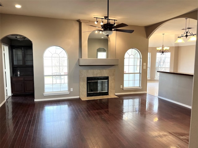 unfurnished living room with baseboards, dark wood finished floors, a tiled fireplace, and ceiling fan with notable chandelier