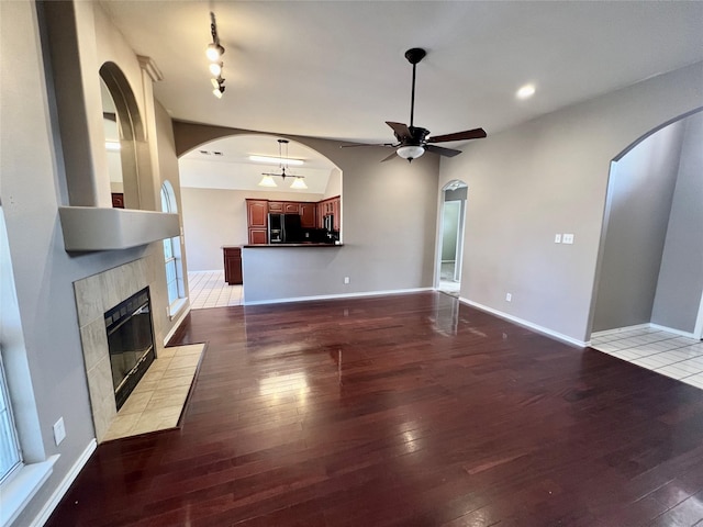 unfurnished living room with baseboards, arched walkways, a ceiling fan, a tiled fireplace, and hardwood / wood-style floors