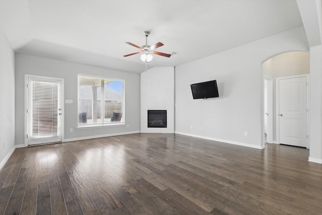 unfurnished living room with a large fireplace, baseboards, dark wood finished floors, arched walkways, and a ceiling fan