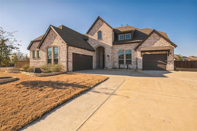 french country inspired facade featuring fence, roof with shingles, an attached garage, concrete driveway, and brick siding