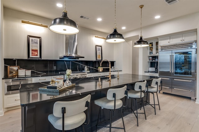 kitchen with light wood-style flooring, appliances with stainless steel finishes, dark countertops, wall chimney exhaust hood, and tasteful backsplash