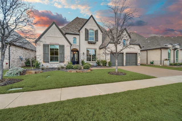 french country style house featuring driveway, an attached garage, a yard, a shingled roof, and brick siding