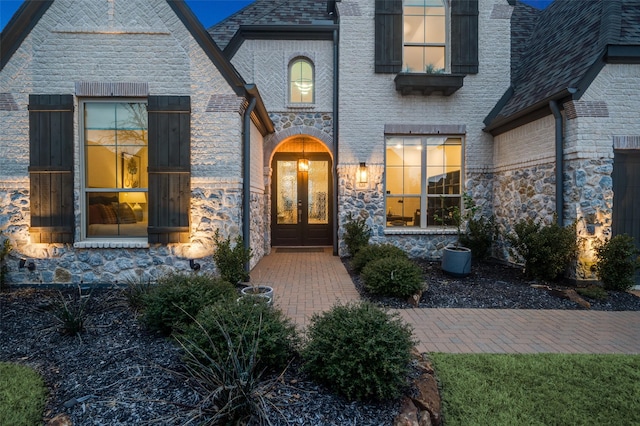 view of exterior entry with brick siding, stone siding, french doors, and roof with shingles