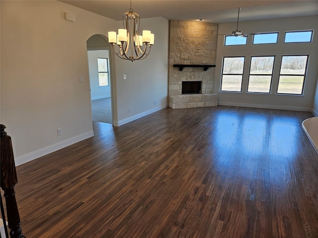 unfurnished living room with arched walkways, dark wood-style flooring, a notable chandelier, a stone fireplace, and baseboards