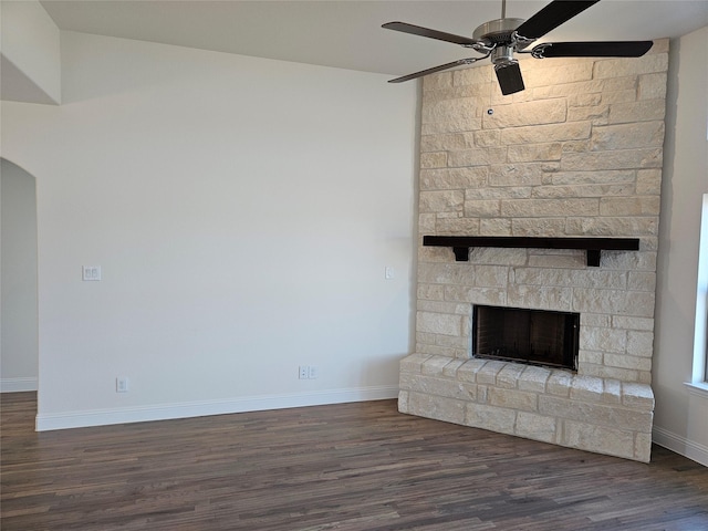 unfurnished living room featuring ceiling fan, arched walkways, a stone fireplace, wood finished floors, and baseboards