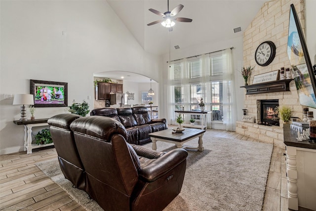 living room with a stone fireplace, visible vents, a ceiling fan, arched walkways, and light wood-style flooring