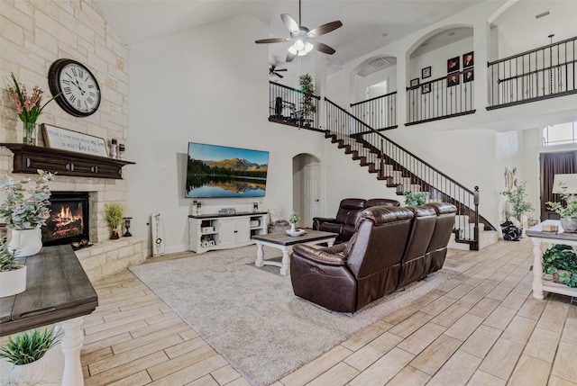 living area featuring arched walkways, a ceiling fan, stairs, a stone fireplace, and wood finish floors