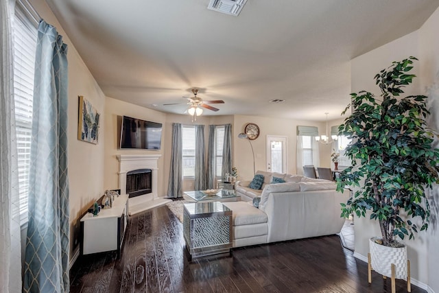 living area with visible vents, a fireplace with raised hearth, baseboards, ceiling fan with notable chandelier, and dark wood-style flooring