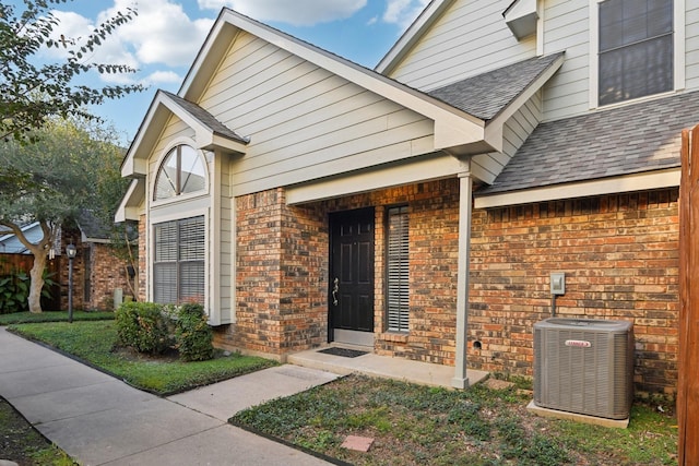 doorway to property with a shingled roof, cooling unit, and brick siding