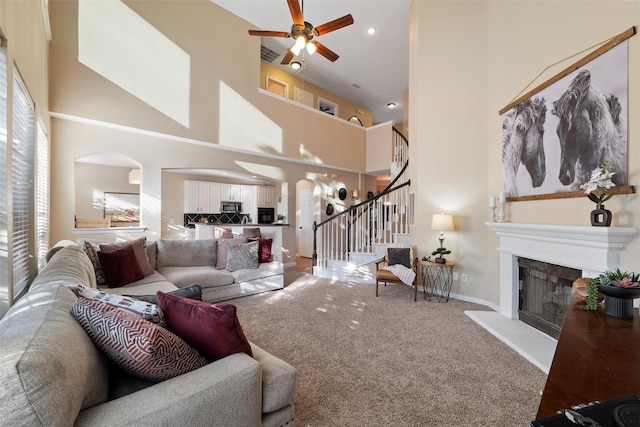 carpeted living area featuring a high ceiling, a glass covered fireplace, ceiling fan, baseboards, and stairs