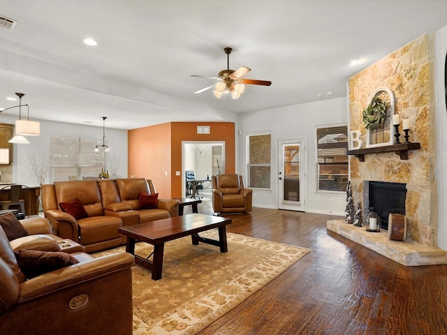 living room featuring dark wood-style floors, a stone fireplace, visible vents, and a ceiling fan