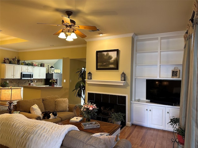 living area with visible vents, a tiled fireplace, ceiling fan, ornamental molding, and wood finished floors