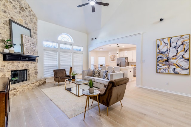 living area featuring light wood-style floors, a stone fireplace, a ceiling fan, a towering ceiling, and arched walkways