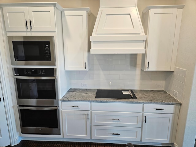 kitchen with black electric stovetop, custom range hood, stainless steel double oven, white cabinets, and built in microwave