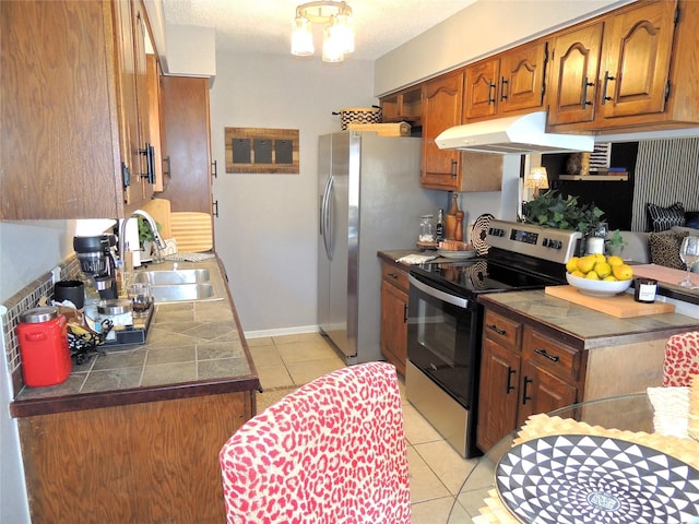 kitchen featuring light tile patterned floors, under cabinet range hood, a sink, appliances with stainless steel finishes, and brown cabinetry