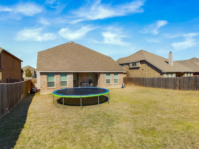 back of house with a fenced backyard, brick siding, roof with shingles, a lawn, and a trampoline