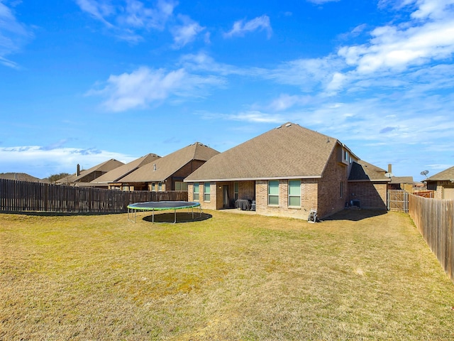 back of property featuring a fenced backyard, brick siding, roof with shingles, a lawn, and a trampoline