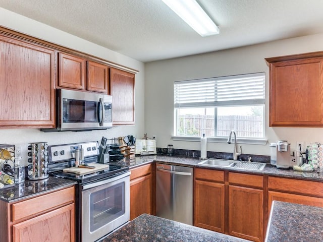 kitchen with brown cabinetry, a textured ceiling, a sink, dark stone counters, and stainless steel appliances