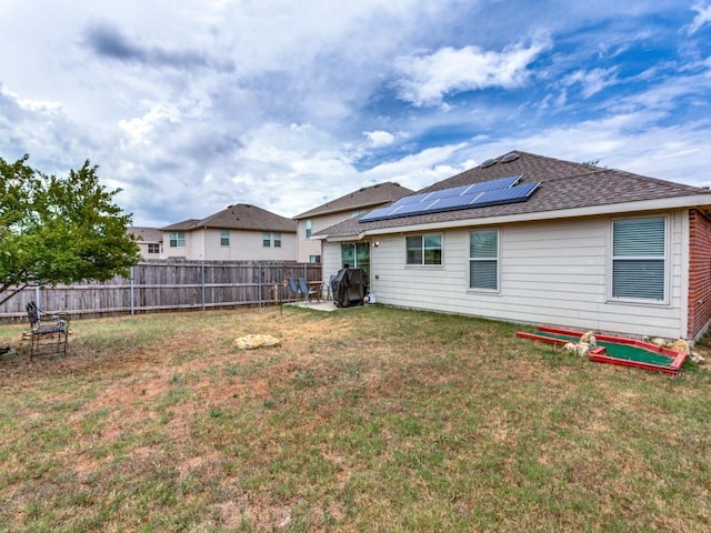 rear view of property with a yard, roof mounted solar panels, a fenced backyard, and roof with shingles