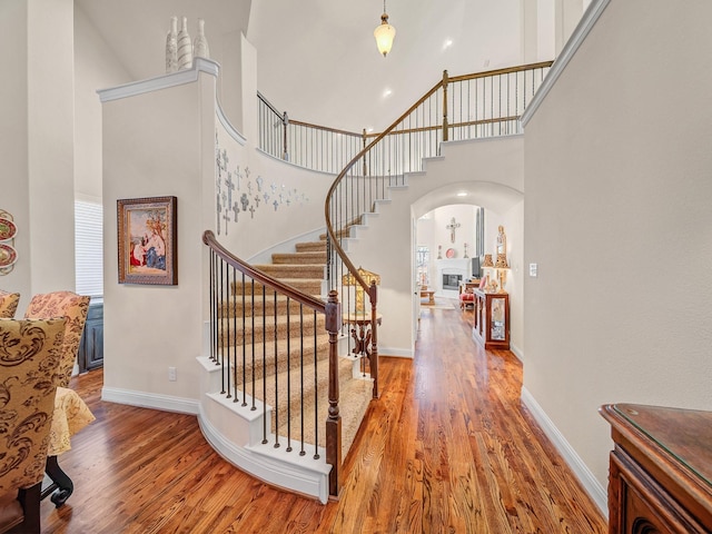 staircase featuring arched walkways, a towering ceiling, a glass covered fireplace, wood finished floors, and baseboards